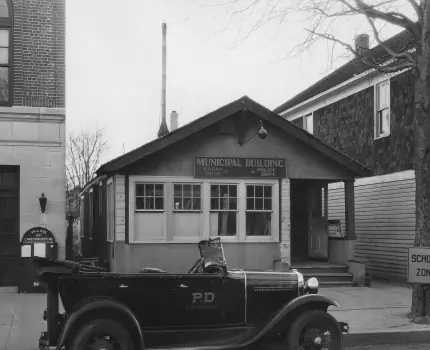 Archival photograph of a municipal building with a vintage police car parked out front